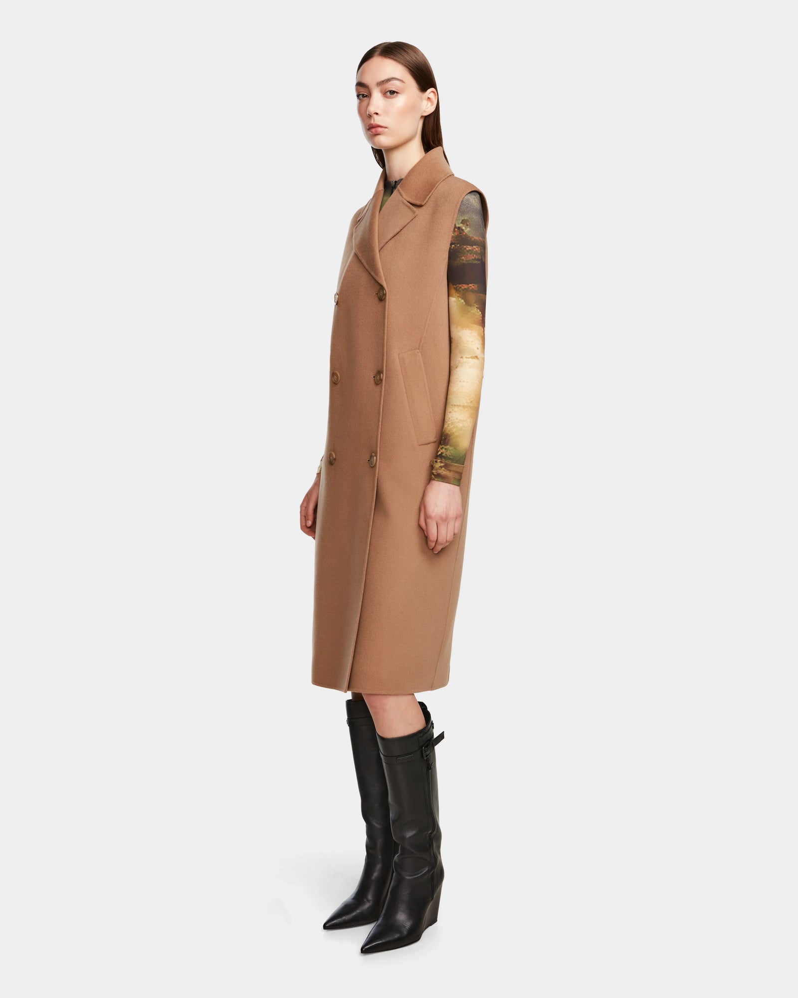 A woman stands sideways against a white background wearing the Rudsak NOURA - 8124698 Camel double-breasted vest styled as a long coat over a printed top and knee-high black boots.