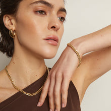 Model. Woman wearing gold jewelry including a necklace, bracelet, and earrings on a neutral background
