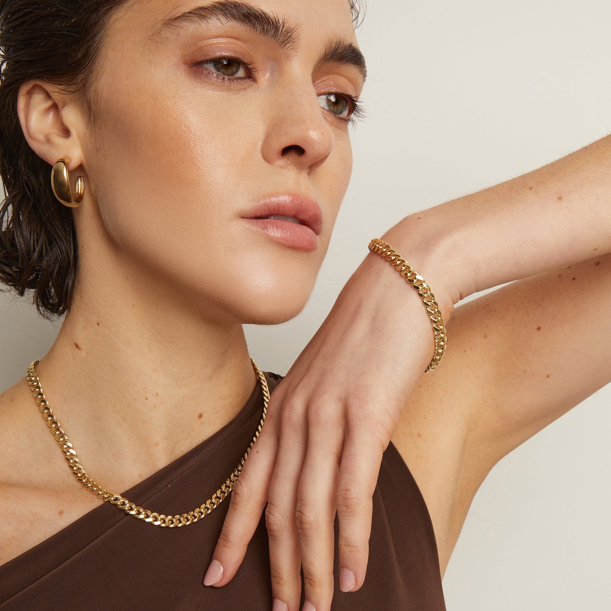 Model. Woman wearing gold jewelry including a necklace, bracelet, and earrings on a neutral background