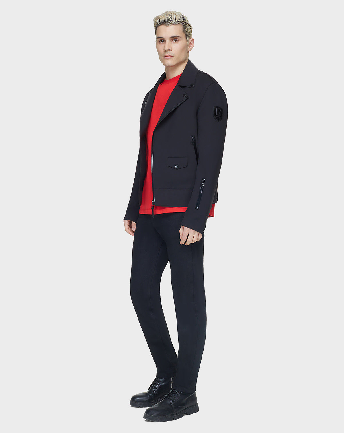 A young man with blond hair, wearing the RUDSAK JUNE - 6122005 BLACK mens jacket over a red shirt, black skinny pants, and black shoes, stands against a plain background facing slightly sideways with a neutral expression.