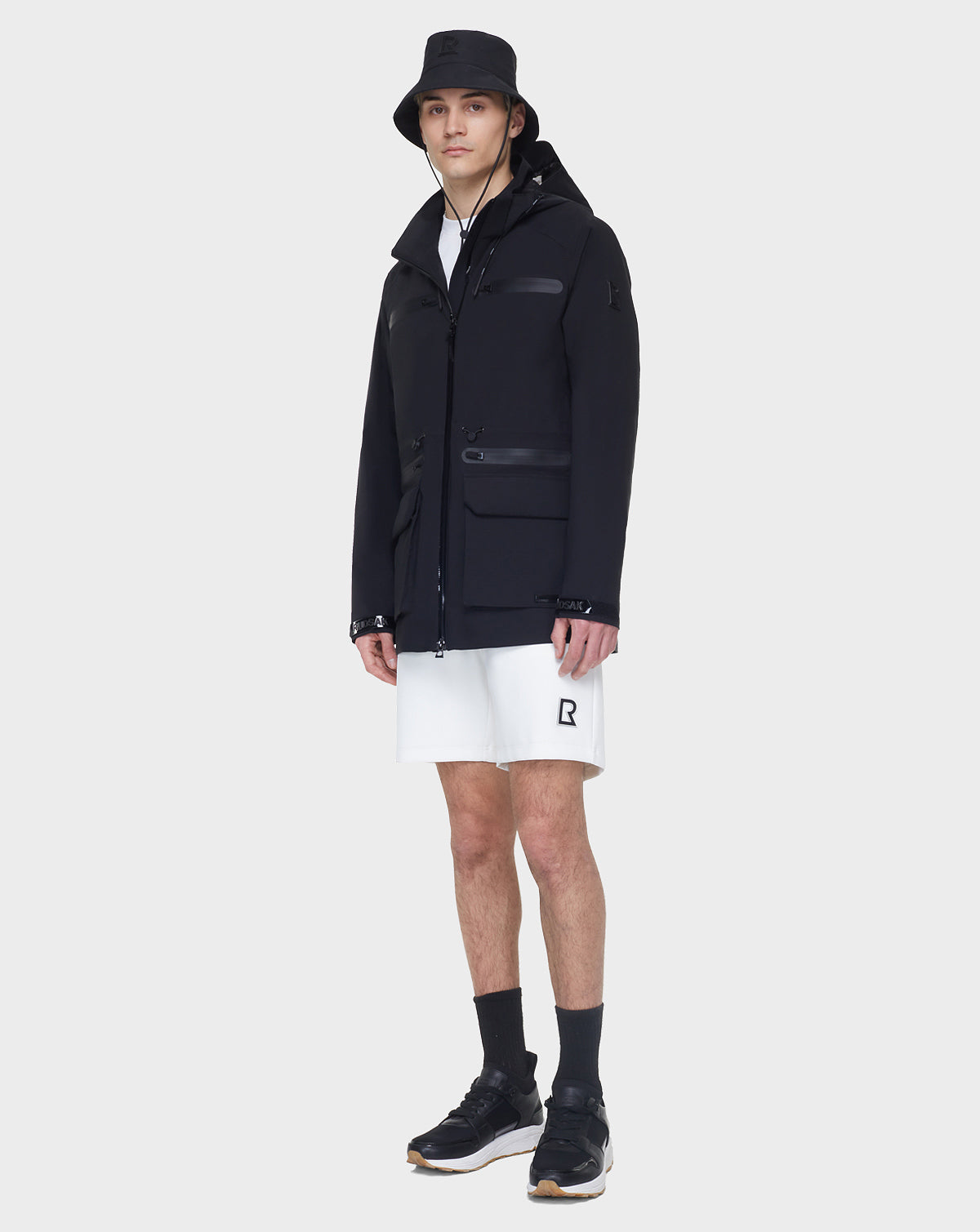A man models the RUDSAK JACY - 6122000 BLACK bucket hat, paired with a mid-length rainwear jacket, white shorts, black socks, and black sneakers, standing against a plain light background with a neutral expression.