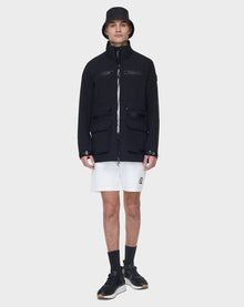 A man faces forward against a white background, wearing the RUDSAK JACY - 6122000 BLACK zip-up rainwear jacket with pockets, black bucket hat, white shorts, black socks, and black sneakers.