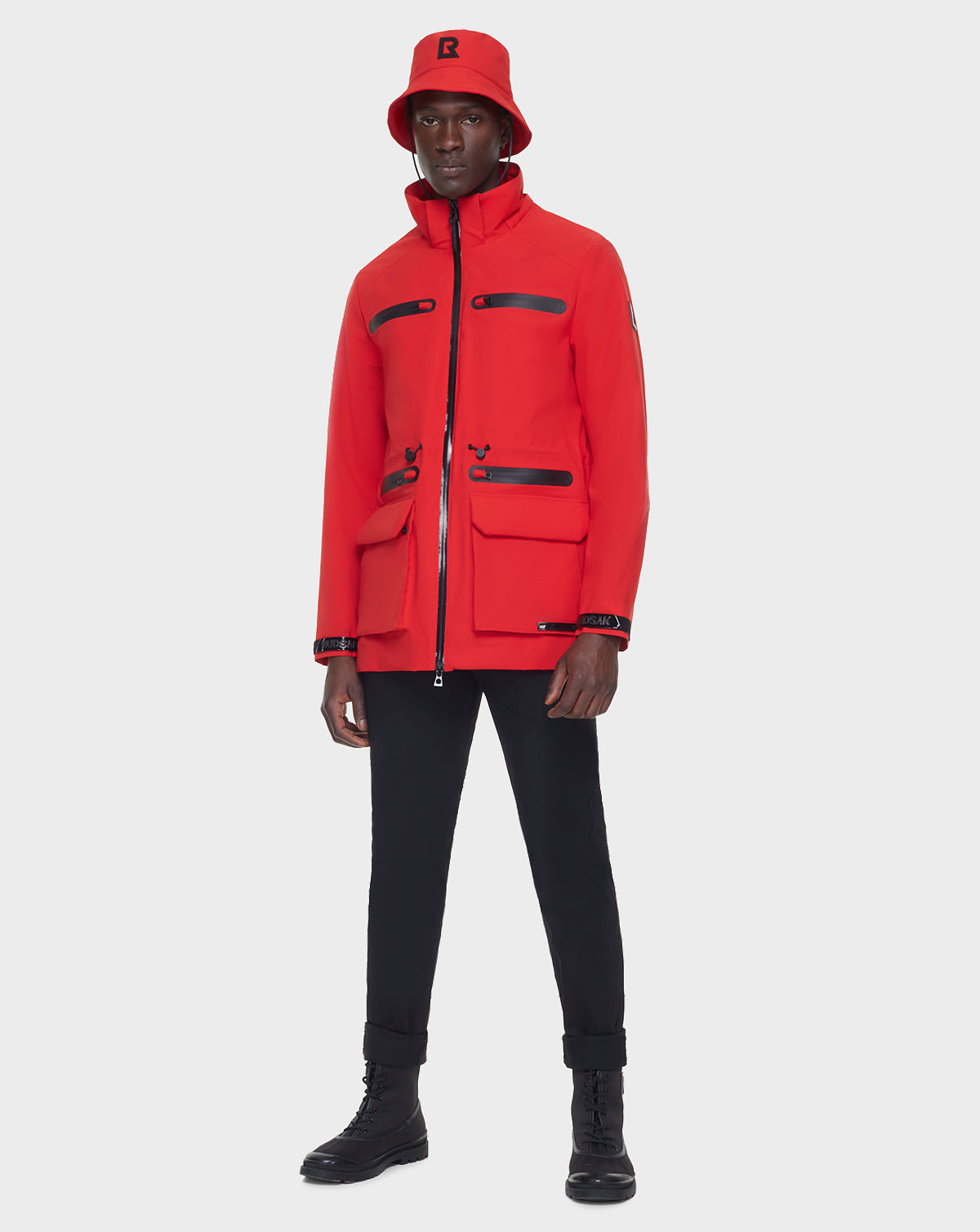 Wearing the RUDSAK JACY - 6122000 POPPY RED raincoat and a matching red bucket hat, paired with black pants and boots, a person stands against a plain white background.