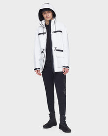 A person wears the RUDSAK JACY - 6122000 WHITE mid-length raincoat, featuring black zippers and pockets, paired with black pants and boots, standing against a plain light gray background.