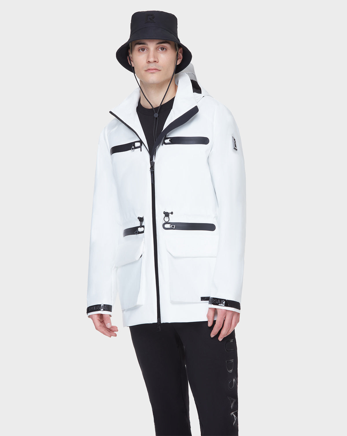 A man wearing the RUDSAK JACY - 6122000 WHITE mid-length raincoat with black zippers and details, black pants, and a black bucket hat stands against a plain light gray background.
