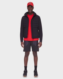 A man faces forward against a plain white background wearing the RUDSAK JAS - 6122006 BLACK men’s windbreaker puffer jacket, a red cap, red t-shirt, black shorts with gray pockets, black socks, and black sneakers.