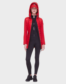 A person stands facing forward wearing the Rudsak MERINA - 8121006 POPPY RED women's light jacket with a hood, a matching red logo cap, black zip-up top, leggings, and sneakers against a plain light background.